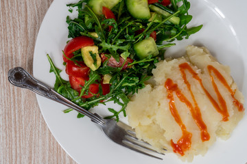 Vegan mashed potatoes on a white plate next to arugula salad with cherry tomatoes and cucumbers. Homemade plant based meal.