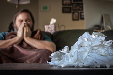 A Caucasian Man Blowing His Nose while Sitting on the Couch and Wearing a Blanket - with a Large Pile of Tissues on the Table in the Foreground