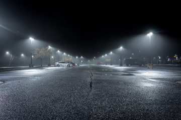 A Lonely Parking Lot on a Cold, Rainy Night - with a Small Collection of Parked Cars in the Background and a Thick Mist Gathering Under the Lights - Powered by Adobe