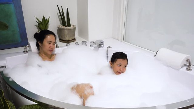 Asian child girl and mom happily bathe in a bathtub filled with white bubbles.
