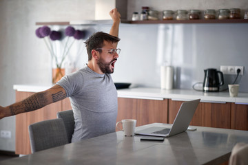 young adult man with beard and tattoo yawns, stretching in front of laptop on table with cup of...