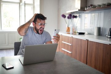 young adult man with beard and tattoo wearing glasses, yawns, looking his cell phone, in front of laptop on table. lifestyle, freelance, social media concept