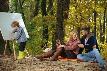 Happy family. Smiling parents look how their kid paints at drawing easel and drink wine. Autumnal vacation. Family picnic at beautiful autumn season forest park. Family relax at nature.
