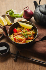 noodles with shrimps and vegetables in bowl near chopsticks, soy sauce, lemon and ginger root, teapot on napkin on wooden table