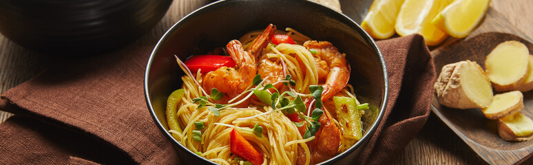 noodles with shrimps and vegetables in bowl near lemon and ginger root on napkin on wooden table,...
