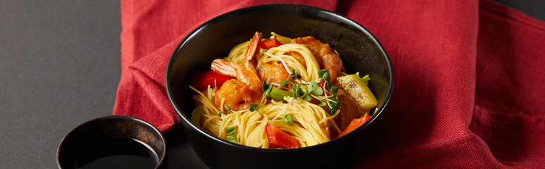 noodles with shrimps and vegetables in bowl near soy sauce on red napkin on black background, panoramic shot