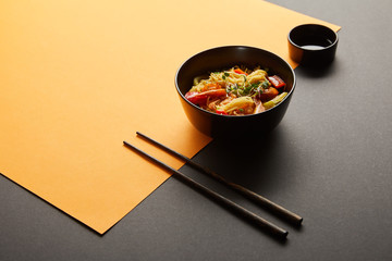 noodles with shrimps and vegetables in bowl near chopsticks, soy sauce on black and yellow background