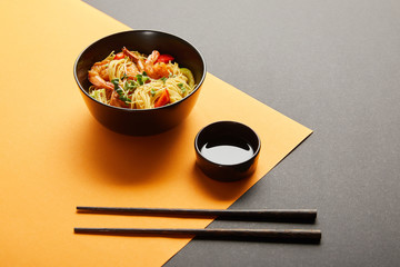 noodles with shrimps and vegetables in bowl near chopsticks, soy sauce on black and yellow background