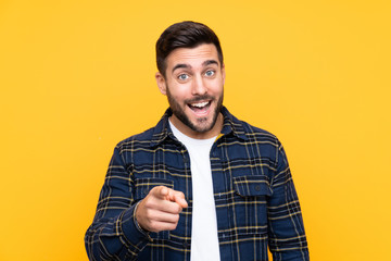Young handsome man with beard over isolated yellow background surprised and pointing front