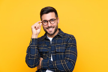 Young handsome man with beard over isolated yellow background with glasses and happy
