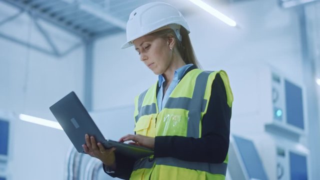 High-Tech Factory: Confident and Professional Female Engineer Wearing Safety Jacket and Hard Hat Holding and Working on Laptop Computer. Modern Bright Industrial Facility. Low Angle Arc Camera Shot