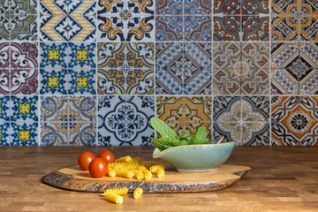 Wooden cuttingboard with small red cherry tomatoes  fusilli pasta and herbs in bowl on wooden kitchencounter with colorful tiles in background