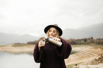 Happy beautiful woman with hat drinking coffee in nature, on a foggy morning Lake and mountains are in the background. She is smiling and poring coffee.