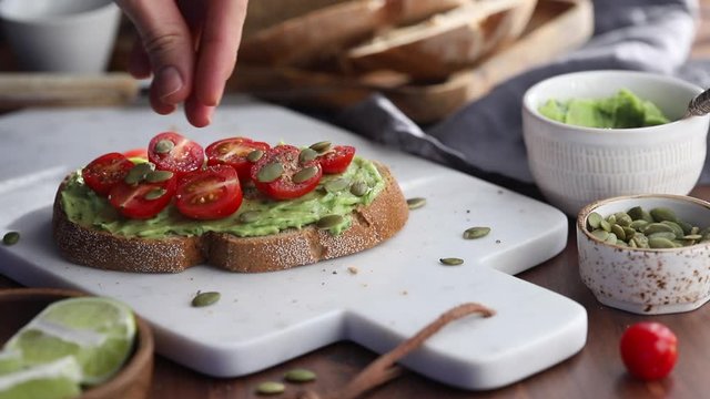 Hand sprinkles pumpkin seeds on an avocado toast with cherry tomatoe