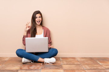 Teenager student girl sitting on the floor with a laptop surprised and showing ok sign
