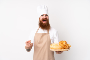 Redhead man in chef uniform. Male baker holding a table with several breads surprised and pointing finger to the side