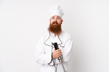 Redhead man using hand blender over isolated white background keeping arms crossed