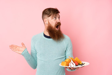 Redhead man with long beard holding waffles over isolated pink background with surprise facial expression