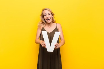 young blonde woman excited, happy, joyful, holding the letter V of the alphabet to form a word or a...