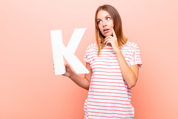 young pretty red head woman confused, doubtful, thinking, holding the letter K of the alphabet to...