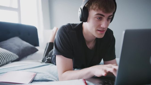 Male College Student Wearing Headphones Works On Bed In Shared House With Laptop 