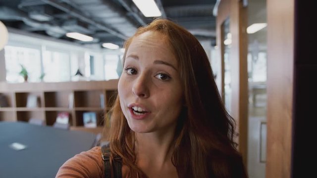 Smiling Casually Dressed Businesswoman Talks Directly To Camera Walking Through Open Plan Office