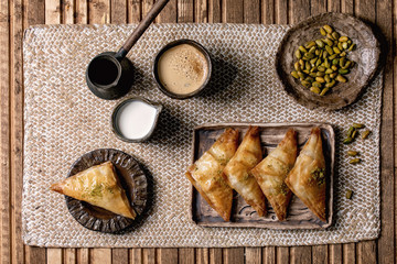 Homemade Turkish traditional dessert baklava with pistachio served on ceramic plate with bowl of nuts, cup of coffee, cream, cezva on straw napkin over wooden background. Flat lay, space