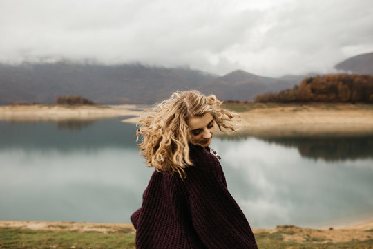 happy girl with curly blond hair dances on a lake alone, her hair is flying because of the wind flow, free as a bird. photo of girl with curly hair standing back to camera over lake background.