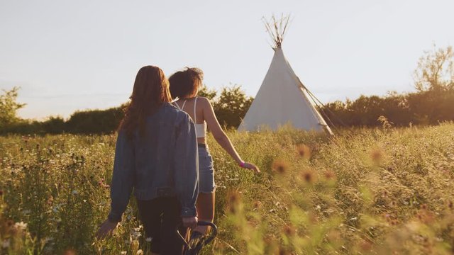 Two Female Friends Walking Pulling Trolley Through Field Towards Teepee On Summer Camping Vacation