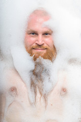 Portrait of a bald man with a long red beard takes a bath with foam. Top view on a cheerful funny guy in soapy water.