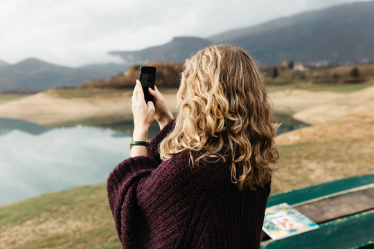 Beautiful woman with curly hair holding mobile phone and taking photos of lake on a cloudy day. She is texting on smartphone and taking selfie. 