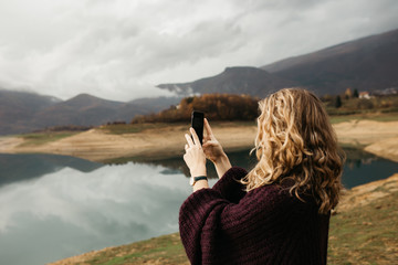 Beautiful woman with curly hair holding mobile phone and taking photos of lake on a cloudy day. She is texting on smartphone and taking selfie. 