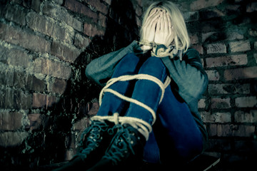 blonde girl with bound hands and feet is sitting in the basement, covering her face with her hands and crying. Concept of violence, kidnapping