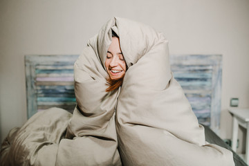 .Pretty young woman smiling just woken up wrapped in her large quilt on a cold winter morning....