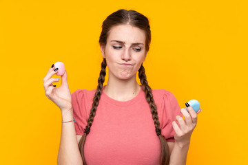 Young woman over isolated yellow background holding colorful French macarons and unhappy