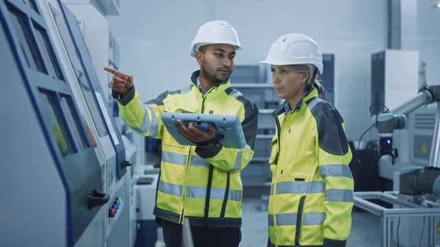 Chief Engineer and Project Manager Wearing Safety Vests and Hard Hats, Use Digital Tablet Controller in Modern Factory, Talking, Optimizing CNC Machinery, Programming Machine for Increasing Efficiency