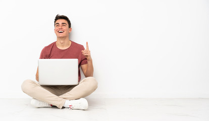 Teenager man sitting on the flor with his laptop pointing up and surprised