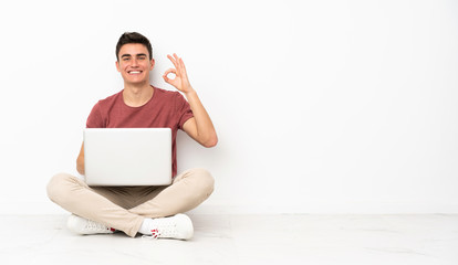 Teenager man sitting on the flor with his laptop showing ok sign with fingers