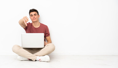 Teenager man sitting on the flor with his laptop showing thumb down with negative expression
