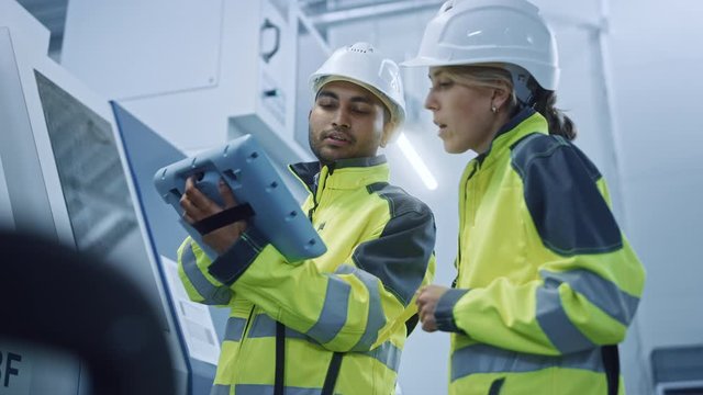 Chief Engineer and Project Manager Wearing Safety Vests and Hard Hats, Use Digital Tablet Controller in Modern Factory, Talking, Programming Machine For Productivity. Low Angle Portraits