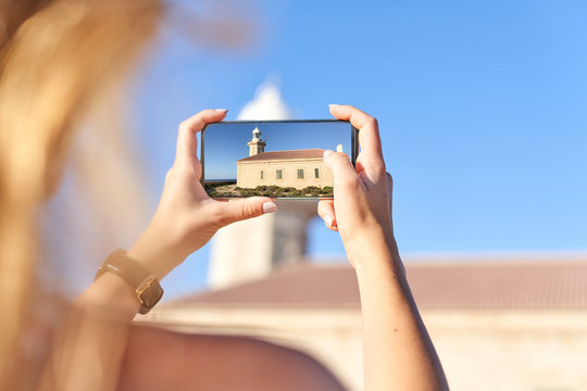 Young woman taking pictures of Punta Nati lighthouse, Menorca, Spain