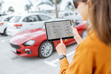 Woman controlling car with mobile application while standing near the vehicle outdoors, close-up view on the tablet with app interface