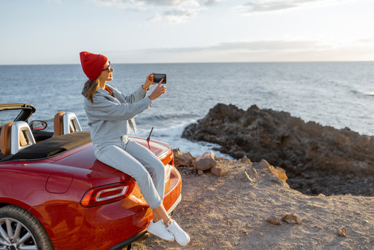 Woman photographing on phone beautiful ocean view, traveling by car on the rocky coastline during a sunset. Carefree lifestyle and car travel concept