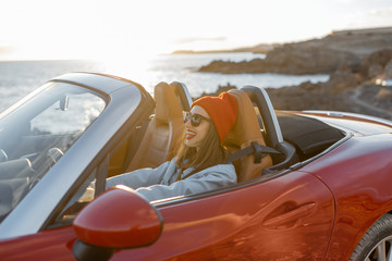 Lifestyle portrait of happy woman in red hat driving convertible car on the rocky ocean coast during a sunset. Carefree lifestyle and car travel concept