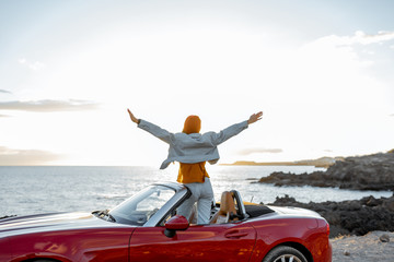 Woman enjoying beautiful view on the ocean, sitting with raised hands on the convertible car during...