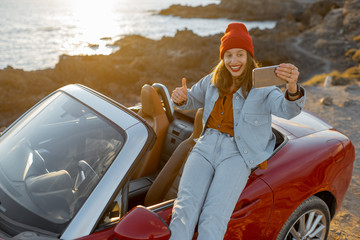 Young stylish woman photographing or vlogging on phone while traveling by car on the rocky coast near the ocean. Lifestyle travel and social influencing concept