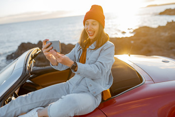 Young stylish woman photographing or vlogging on phone while traveling by car on the rocky coast...