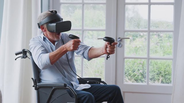 Senior Disabled Man In Wheelchair At Home Wearing Virtual Reality Headset Holding Gaming Controllers