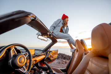 Woman enjoying beautiful view on the ocean, sitting on the car roof top during a sunset. Nature enjoyment and carefree travel by car concept