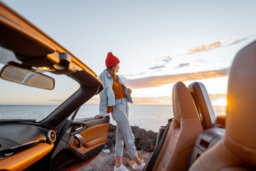 Naklejka premium Woman enjoying beautiful view on the ocean, standing on the rocky coast during a sunset, view from the vehicle interior. Nature enjoyment and car travel concept
